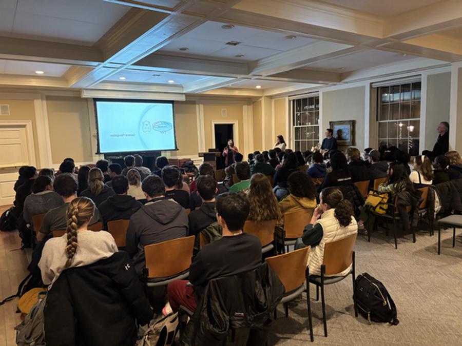 A woman stands in front of a room describing what is being projected on screen, addressing a room full of students seated in chairs.